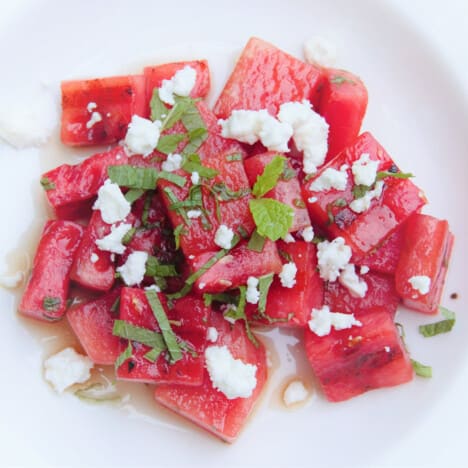 Looking down onto a white plate with grilled watermelon cubes garnished with crumbled goat cheese and sliced mint.