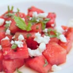 A close up of grilled, cubed watermelon on a white plate, garnished with crumbled white cheese and sliced mint.