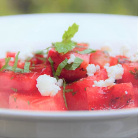 A close up of grilled, cubed watermelon on a plate with crumbled goat cheese and fresh mint.