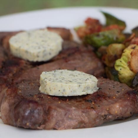Two steaks each topped with a pat of fresh herb butter on a dinner plate.