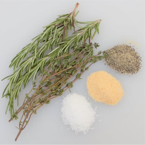 A white chopping board with sprigs of thyme and rosemary next to piles of salt, garlic, and pepper.