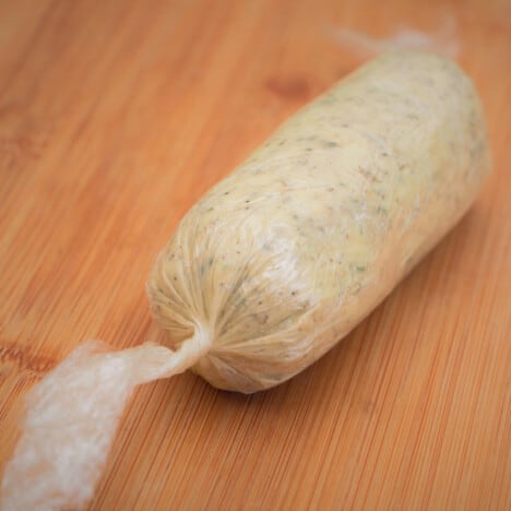 A chopping board with a plastic wrapped roll of herb butter sitting on it.