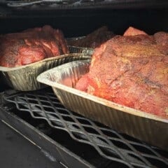 Two pork butts (shoulders) cooking in trays in a smoker