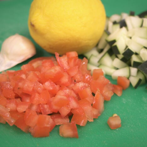 A green chopping board with diced tomato and cucumber, a lemon and clove of garlic.