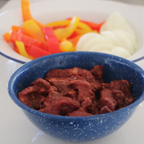 A blue camp bowl filled with seasoned sliced meat and raw bell peppers and onions in the background.