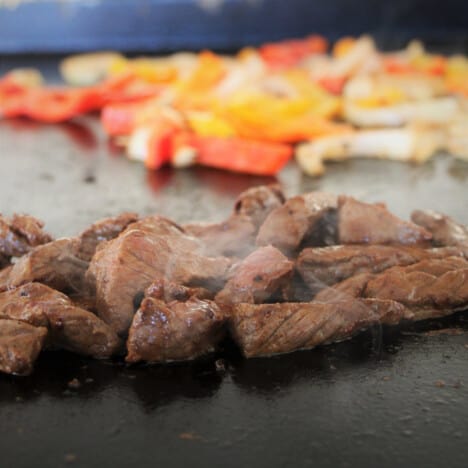 A flat top grill with meat cooking in the front and bell peppers and onions towards the back.