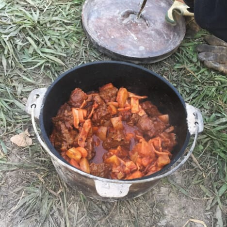 A Dutch oven sitting on the grass filled with campfire beef stew and the lid sitting off to the side.