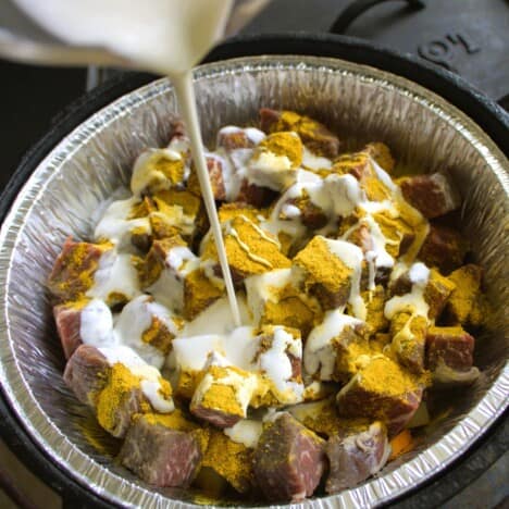 Cream being poured over a lined Dutch oven filled with cubed beef and vegetables.