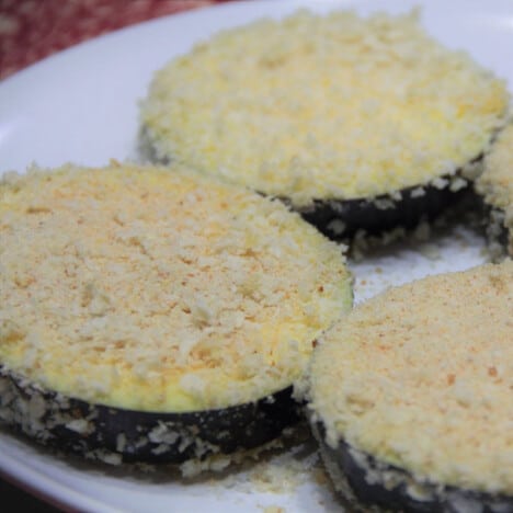 Breaded eggplant slices sit on a plate waiting to be fried.