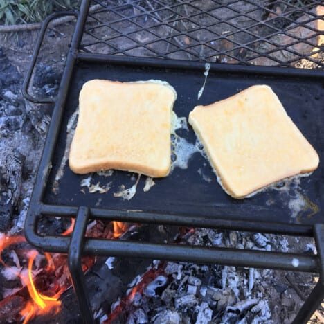 Two slices of raw French toast cooking on a skillet over coals.