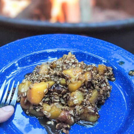 A blue camp plate with a serving of peach crisp with a campfire in the background.