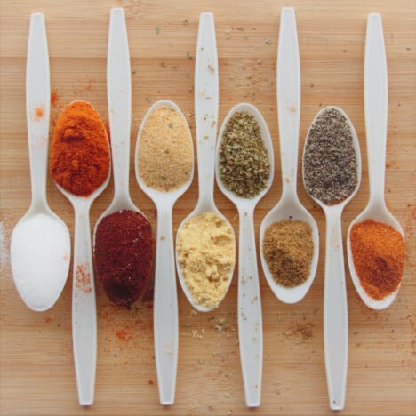 Looking down on white spoons with a range of spices in them sitting on a wooden chopping board.