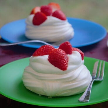Two individual pavlovas with cream and berries sit on colorful camping plates.