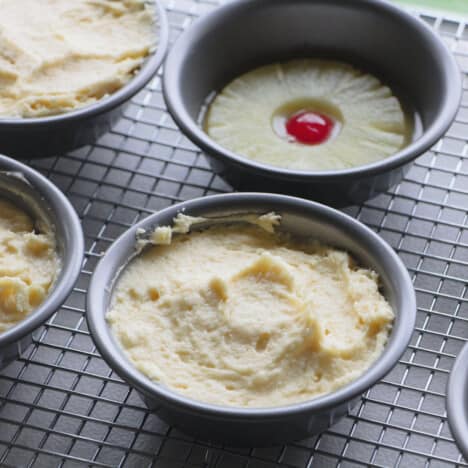 A mini round cake tin with batter in it with one behind still only with the pineapple and cherry.