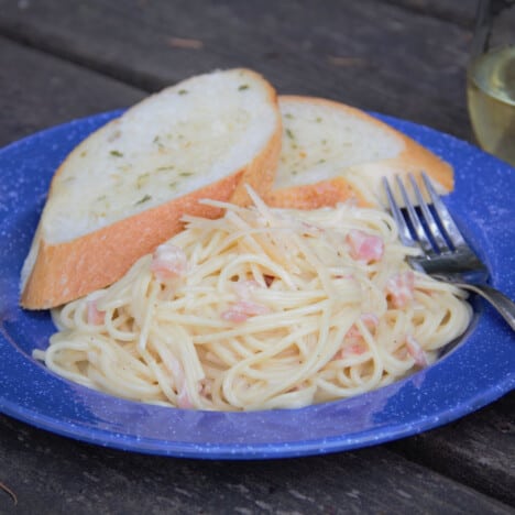 A blue camp plate on a picnic table with a serving of pasta carbonara and two slices of garlic bread.
