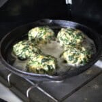 A skillet on a gas camp stove frying five spinach patties.