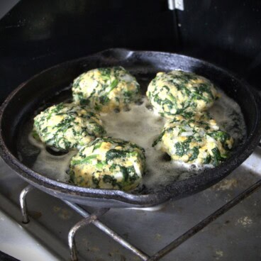 A skillet on a gas camp stove frying five spinach patties.