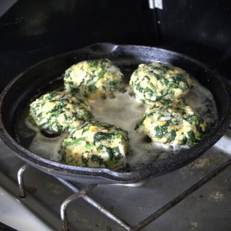 A skillet on a gas camp stove frying five spinach patties.