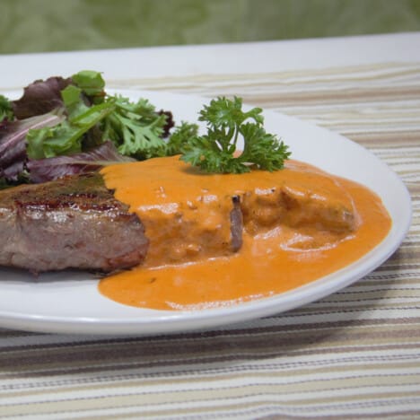 A steak sits on a white plate smothered in red steak Diane sauce.