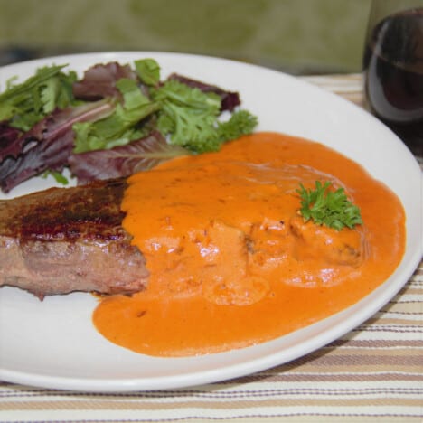 Looking down on a white plate with a steak covered with red steak Diane sauce and a side salad.