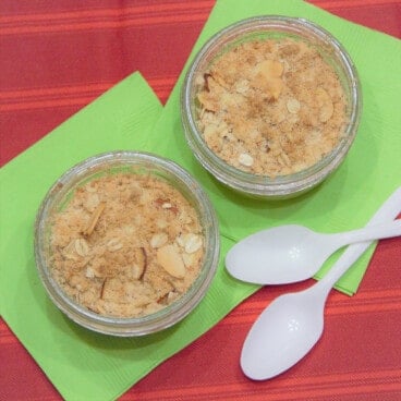 Looking down on two apple crisps in small canning jars on a green napkins on a red table.
