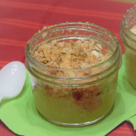 A small glass canning jar filled with apple crisp, on a green napkin with a plastic spoon.