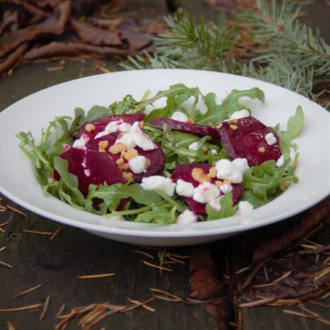 A large shallow white bowl holds a salad of arugula, sliced beets, goat cheese, and walnuts.