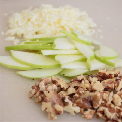 A white chopping board with piles of white cheese, sliced apple, and chopped walnuts.