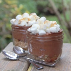 Two small glass canning jars filled with chocolate mousse and topped with toasted mini marshmallows sit on a picnic bench with two spoons.