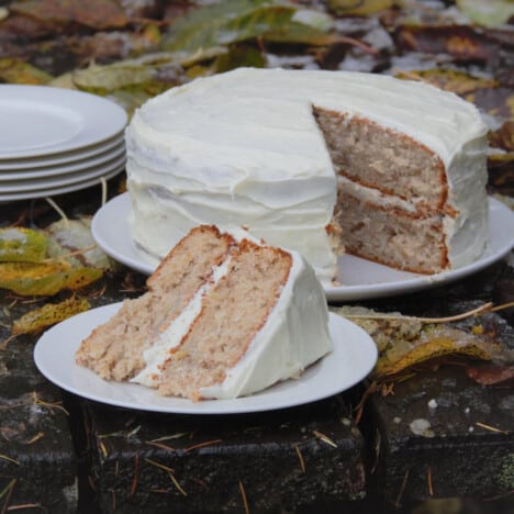 A slice of hummingbird cake is removed and on a plate, in front of the remaining whole cake.