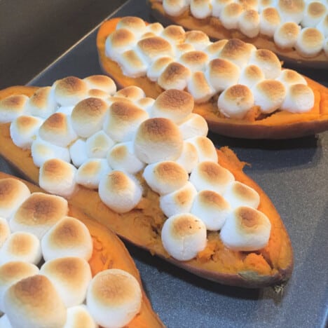 A line of stuffed sweet potatoes covered in browned mini marshmallows sit on a baking sheet.