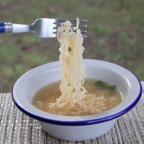 Noodles being lifted from a camp bowl of soup with a fork.