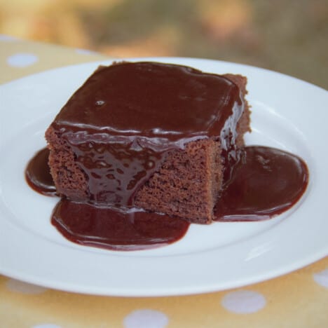 A square slice of chocolate cake served on a white plate topped with a chocolate sauce.