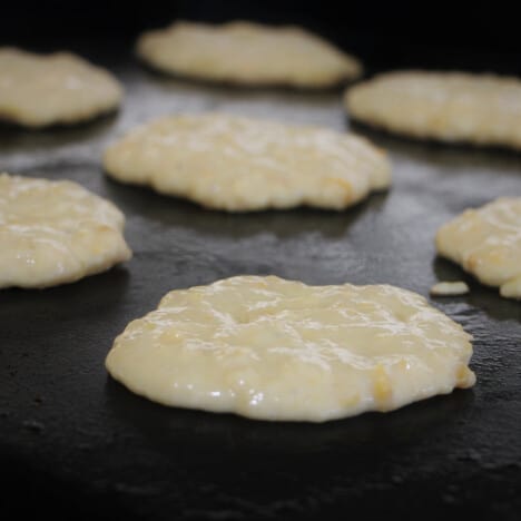 Raw corn fritters cooking on a flat top grill.