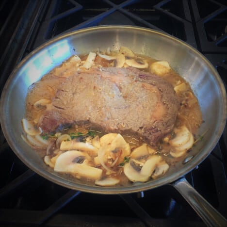 A braised steak sits in a saute pan along with sliced mushrooms.