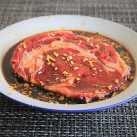 A camp plate with a steak marinating in it, the marinade is dark in color with finely diced ginger and garlic visible.