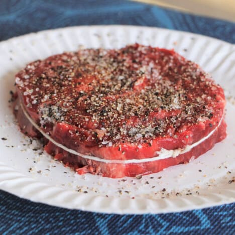 A butcher twine tied raw ribeye steak covered with a course steak rub resting on a paper plate.