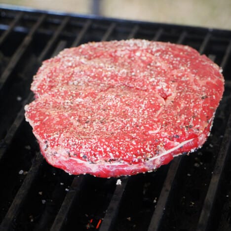 A ribeye steak rounded with a tied piece of butchering string, on a set of grill grates with a light coating of rub.