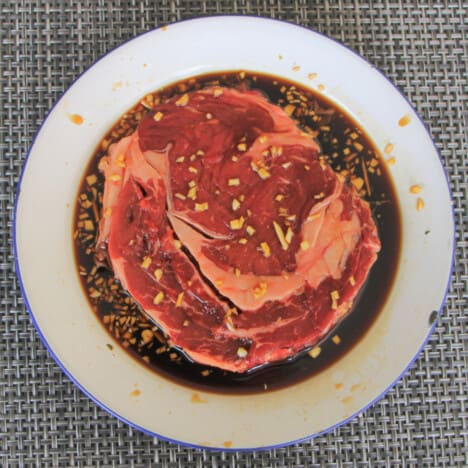 A white camp plate with a ribeye steak sitting in a dark marinade with lots of finely diced garlic and ginger.