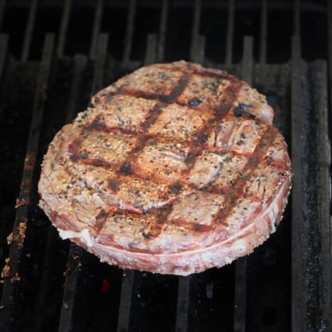 A round tied steak with rub cooking on grill grates with a perfect cross hatch of grill marks.