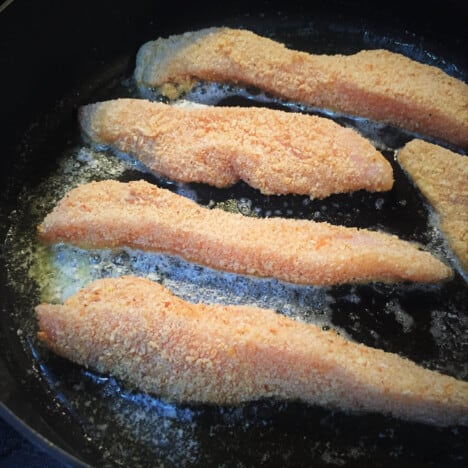 Raw crumbed chicken tenders frying in a skillet.