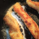 Looking down into a skillet with golden brown chicken tenders sizzling in the oil.