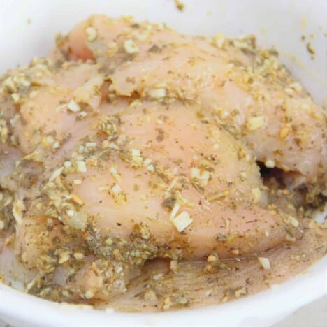 A white bowl full of raw chicken breast being marinated in a marinade.