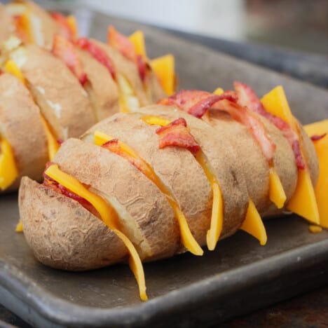 Two baked potatoes cut in a hasselback fashion with slices of cheese and bacon in the slits.