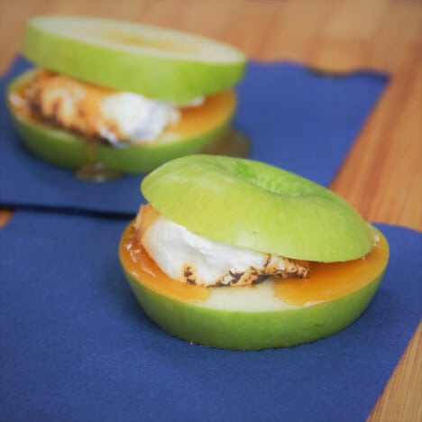 Two apple smores, with marshmallow and caramel sauce, on blue napkins.