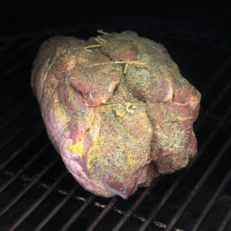 A raw leg of lamb prepared with a mustard slather and rub, as well as being trussed is sitting on a grill grate.