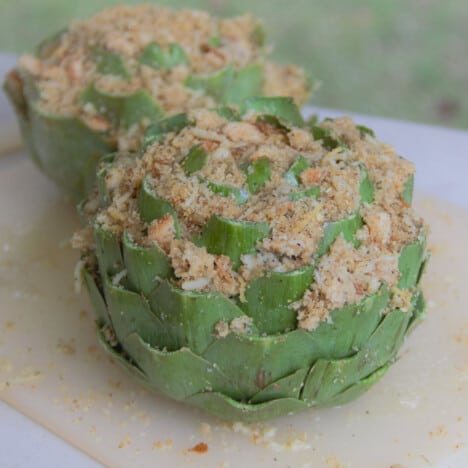 A raw stuffed artichoke, with breadcrumbs popping out, sits on a white cutting board.