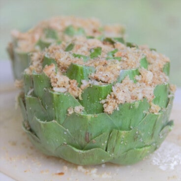 A close up of a raw stuffed artichoke, with breadcrumbs spilling out.