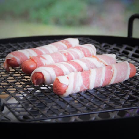 Four hot dogs are wrapped in raw bacon and sitting on a grill grate.