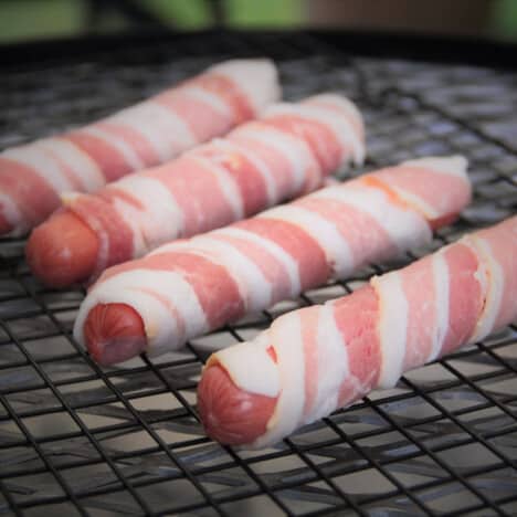Four hot dogs wrapped with raw bacon sit on a smoking grate.
